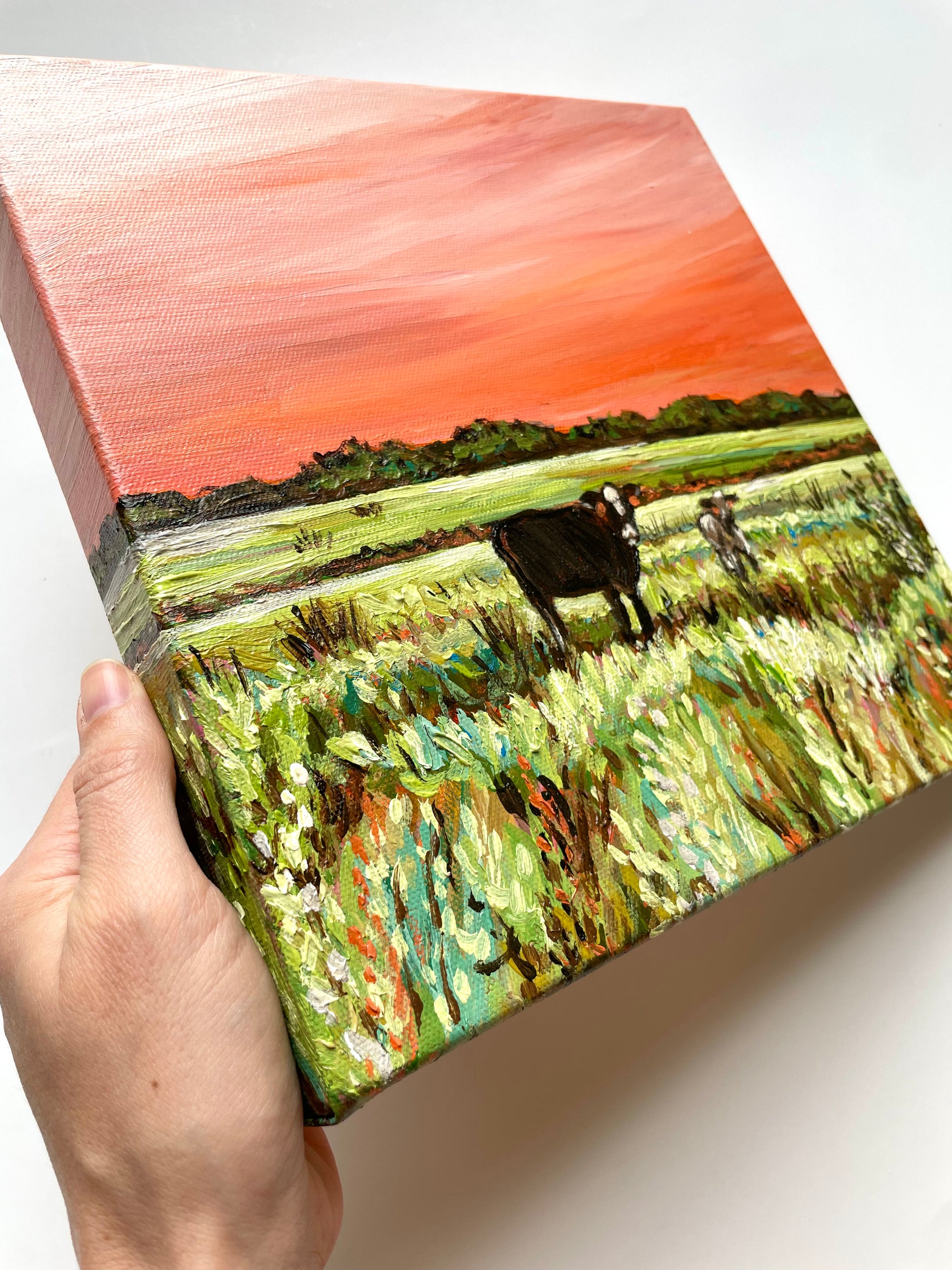 Hand holding a small painting of a landscape with cows.