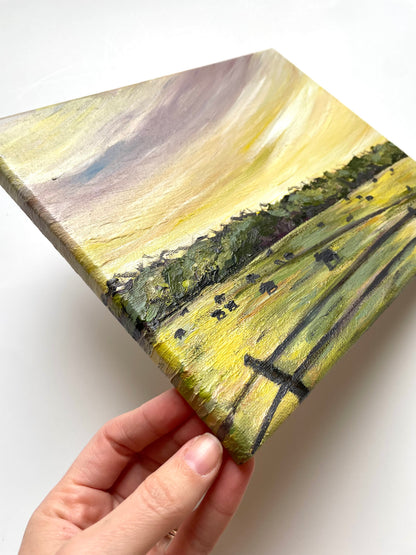 Hand holding a small rectangular painting of a landscape with fields and trees and purple and yellow sunset 