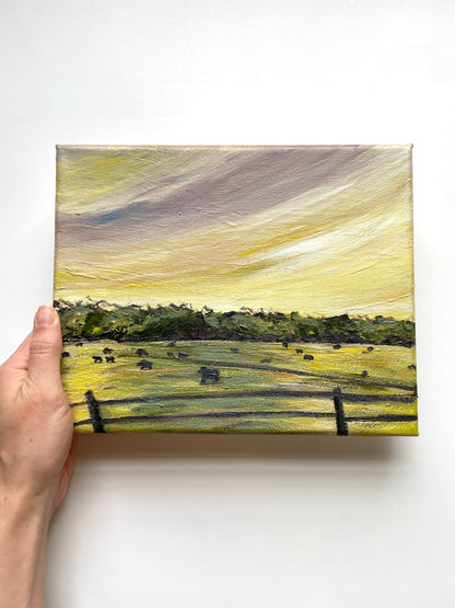 Hand holding a small landscape painting with a field and fence_purple and yellow sunset_cows in field